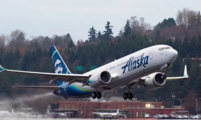 Alaska Airlines plane lifting off from a runway with a forested backdrop behind it.