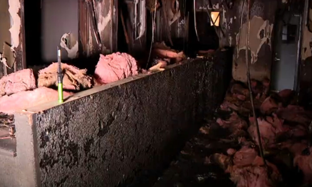 Charred interior with pink insulation on a ledge, burned walls, and debris on the floor near a green tool.