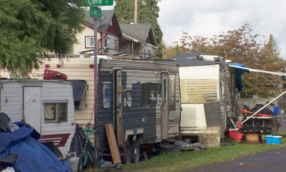 Row of weathered campers and scattered junk in a residential yard beside a street corner.",