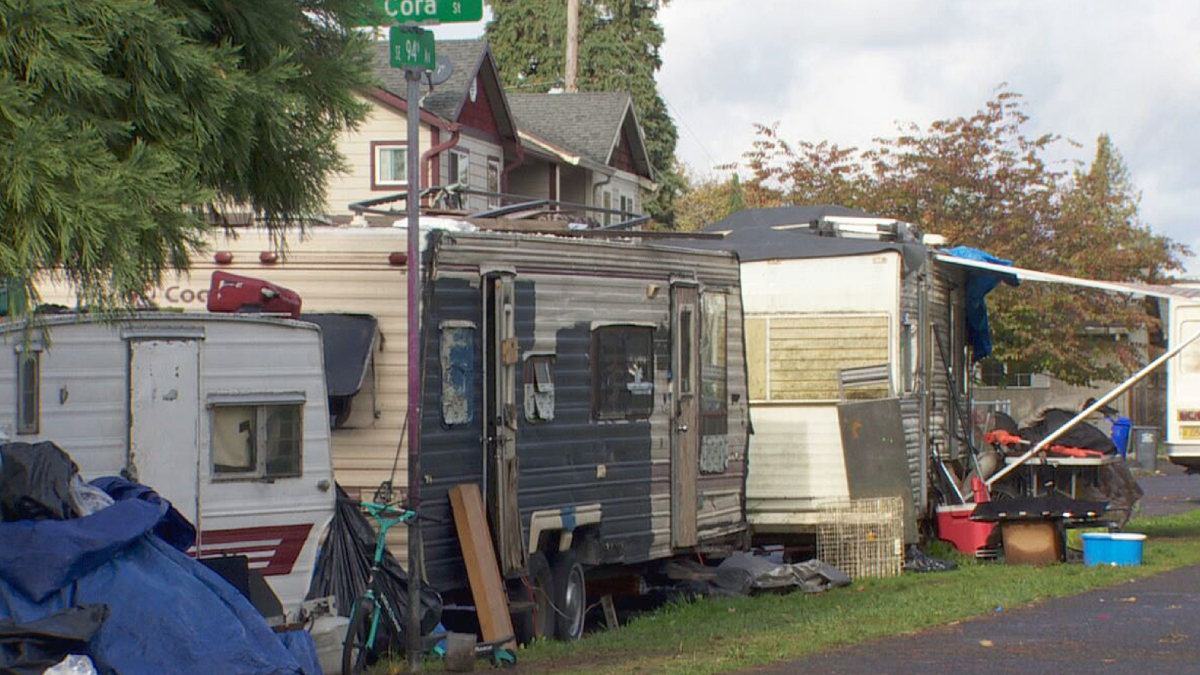 Row of weathered campers and scattered junk in a residential yard beside a street corner.",