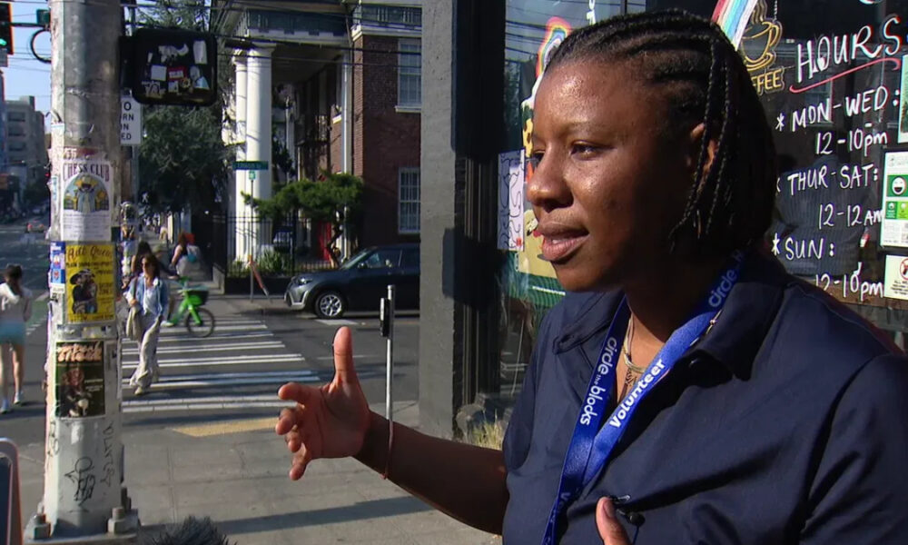Black woman in a navy shirt with a 'Volunteer' lanyard speaking and gesturing on a city sidewalk, with posters on a pole behind her.