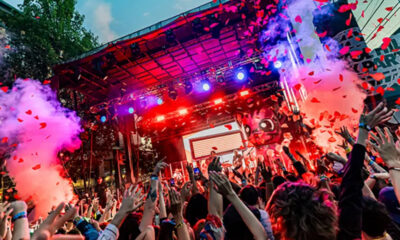 Crowd at a vibrant outdoor concert with raised hands, colorful stage lights, and pink smoke.