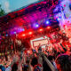 Crowd at a vibrant outdoor concert with raised hands, colorful stage lights, and pink smoke.