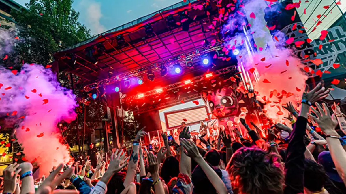 Crowd at a vibrant outdoor concert with raised hands, colorful stage lights, and pink smoke.
