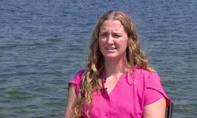 Woman in a bright pink blouse sits by a lake, being interviewed outdoors, with a small lapel microphone clipped to her shirt.