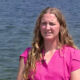 Woman in a bright pink blouse sits by a lake, being interviewed outdoors, with a small lapel microphone clipped to her shirt.