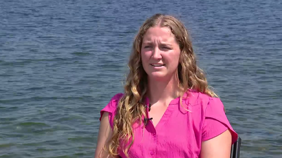 Woman in a bright pink blouse sits by a lake, being interviewed outdoors, with a small lapel microphone clipped to her shirt.