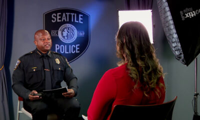 Police officer in uniform being interviewed in a studio, with a Seattle Police shield backdrop visible behind him and a reporter facing away in a red top.