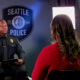 Police officer in uniform being interviewed in a studio, with a Seattle Police shield backdrop visible behind him and a reporter facing away in a red top.