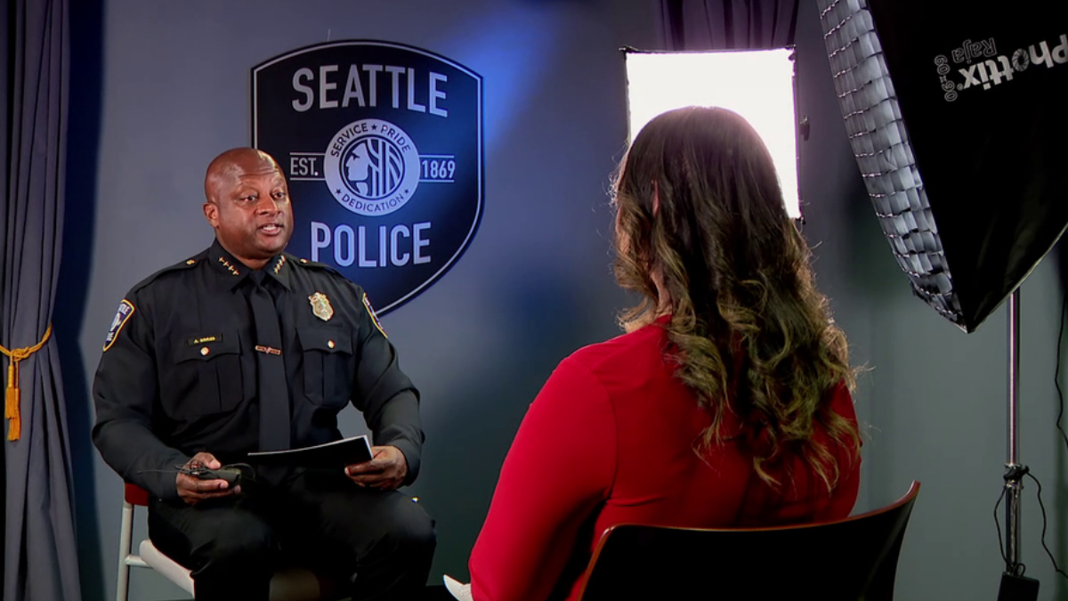 Police officer in uniform being interviewed in a studio, with a Seattle Police shield backdrop visible behind him and a reporter facing away in a red top.