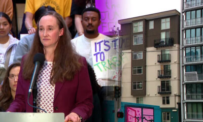 Woman in a maroon blazer speaks at a podium with a microphone, addressing an audience of supporters behind her; urban street scene to the right.