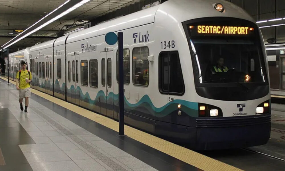 White light rail train at an airport-bound station, displaying SEATAC/AIRPORT with teal wave graphics along the side.