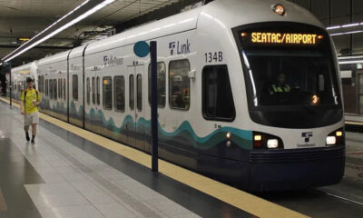 White light rail train at an airport-bound station, displaying SEATAC/AIRPORT with teal wave graphics along the side.