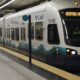 White light rail train at an airport-bound station, displaying SEATAC/AIRPORT with teal wave graphics along the side.