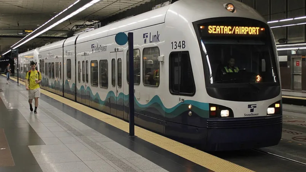 White light rail train at an airport-bound station, displaying SEATAC/AIRPORT with teal wave graphics along the side.