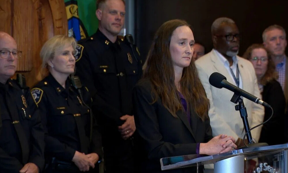 Woman in a dark blazer speaks at a podium with a microphone, flanked by police officers and officials at a press conference.