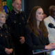 Woman in a dark blazer speaks at a podium with a microphone, flanked by police officers and officials at a press conference.