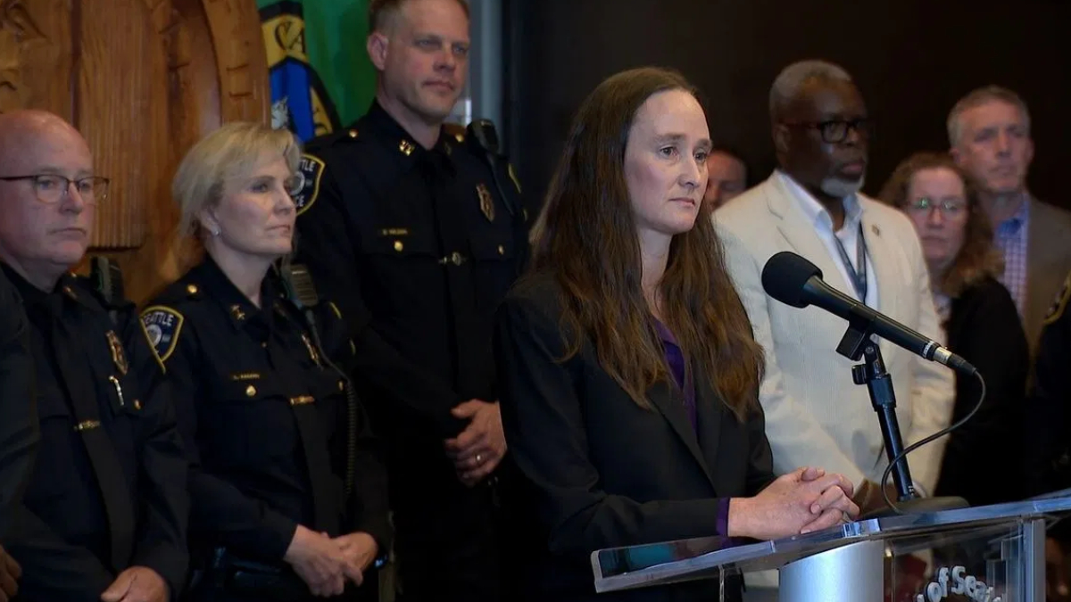 Woman in a dark blazer speaks at a podium with a microphone, flanked by police officers and officials at a press conference.