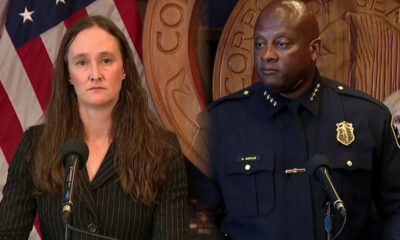 Woman in a pinstripe suit speaks at a podium microphone, flanked by a uniformed police officer, with U.S. flag and government seal in the background.