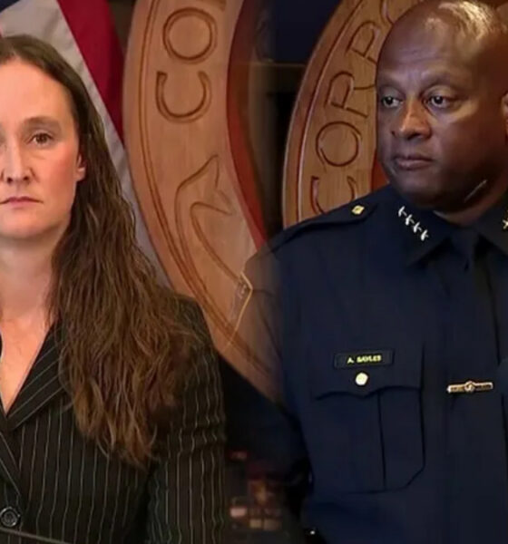 Woman in a pinstripe suit speaks at a podium microphone, flanked by a uniformed police officer, with U.S. flag and government seal in the background.