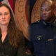 Woman in a pinstripe suit speaks at a podium microphone, flanked by a uniformed police officer, with U.S. flag and government seal in the background.