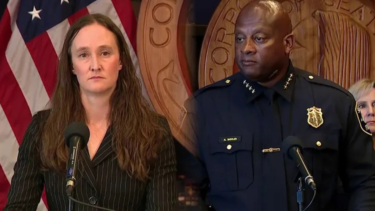 Woman in a pinstripe suit speaks at a podium microphone, flanked by a uniformed police officer, with U.S. flag and government seal in the background.