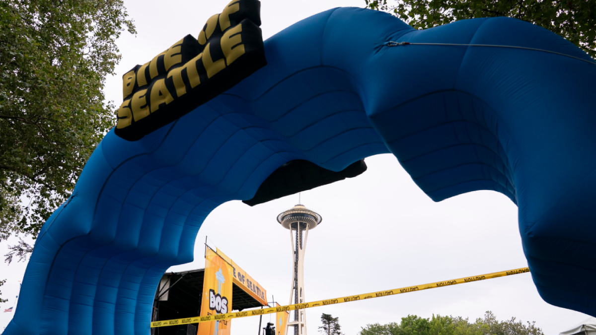 Large blue inflatable arch forms a finish line gate at an outdoor Seattle event, with the Space Needle visible in the background and caution tape across the area.
