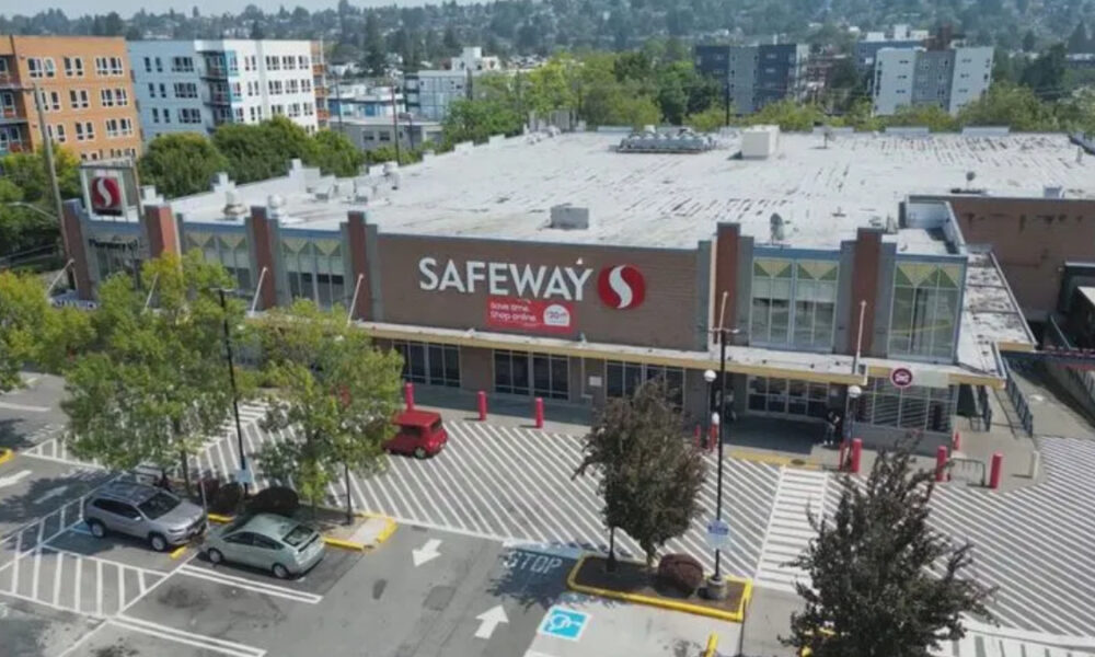 Aerial view of a Safeway supermarket with large front sign, surrounding parking lot, and striped pedestrian crosswalks outside the entrance.