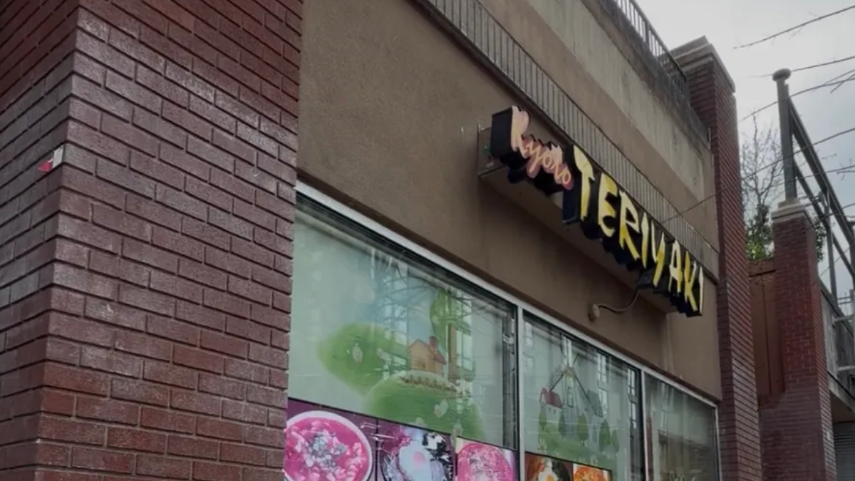 Exterior view of a brick storefront with a sign that appears to say 'Teriyaki' above large display windows with colorful food posters inside.