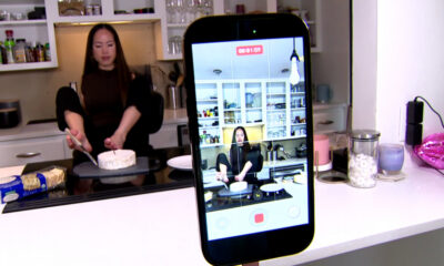 Woman in a kitchen decorates a cake on a countertop while a smartphone on a stand records the scene.