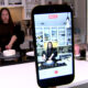 Woman in a kitchen decorates a cake on a countertop while a smartphone on a stand records the scene.