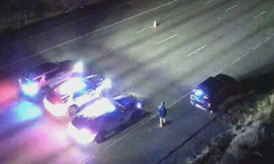 Nighttime highway crash scene with several damaged vehicles and blue/red emergency lights flashing; a person in a blue jacket stands near the center of the road.