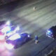 Nighttime highway crash scene with several damaged vehicles and blue/red emergency lights flashing; a person in a blue jacket stands near the center of the road.