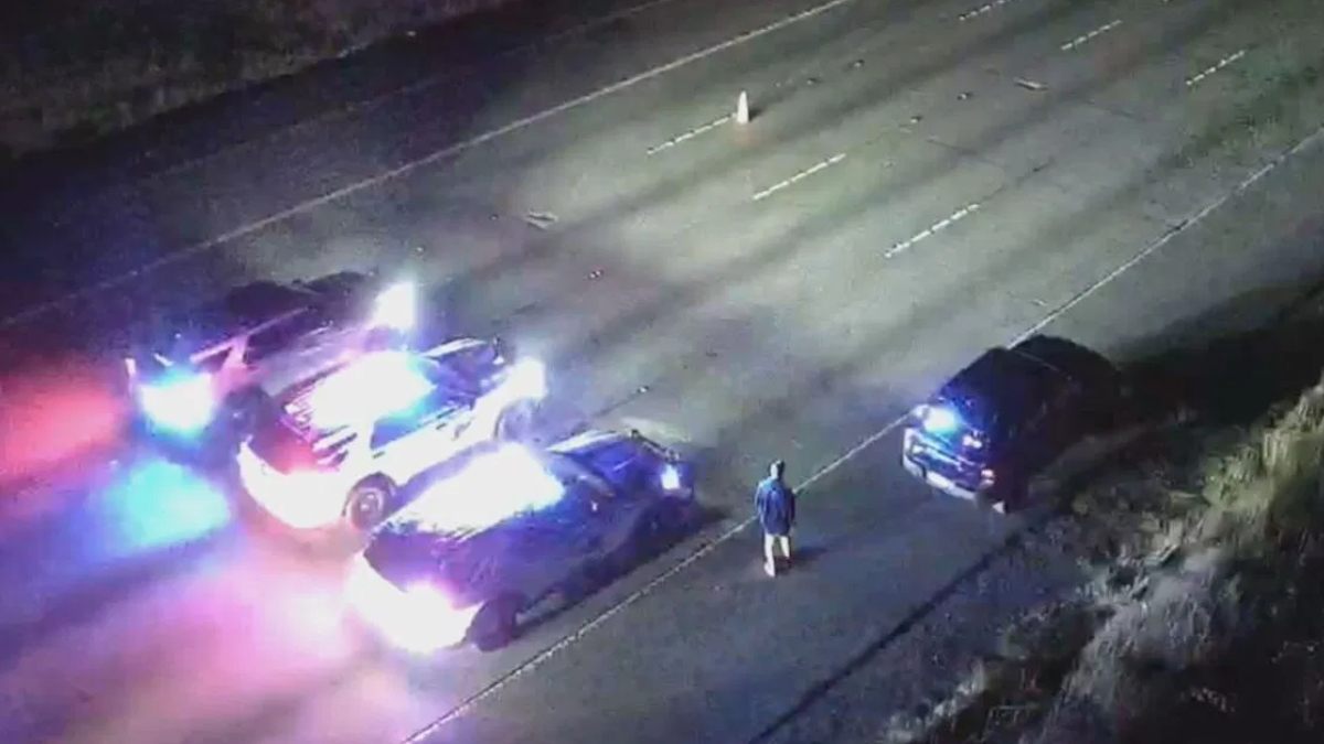 Nighttime highway crash scene with several damaged vehicles and blue/red emergency lights flashing; a person in a blue jacket stands near the center of the road.
