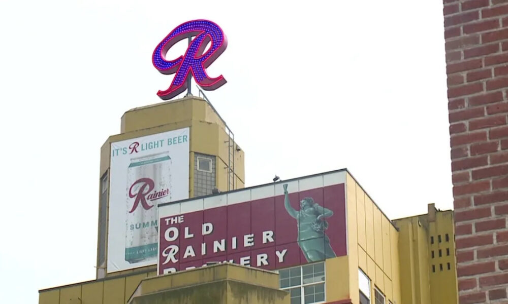 Neon red R sign atop a retro Rainier beer building with banners reading 'The Old Rainier Brewery' and 'It's R Light Beer'.