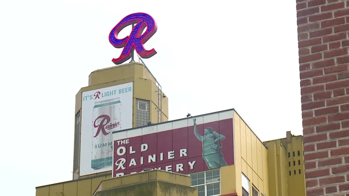Neon red R sign atop a retro Rainier beer building with banners reading 'The Old Rainier Brewery' and 'It's R Light Beer'.