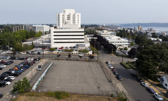 Aerial view of a large white multi-story hospital-like building with surrounding parking lots and streets near a waterfront cityscape.
