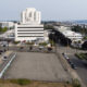 Aerial view of a large white multi-story hospital-like building with surrounding parking lots and streets near a waterfront cityscape.