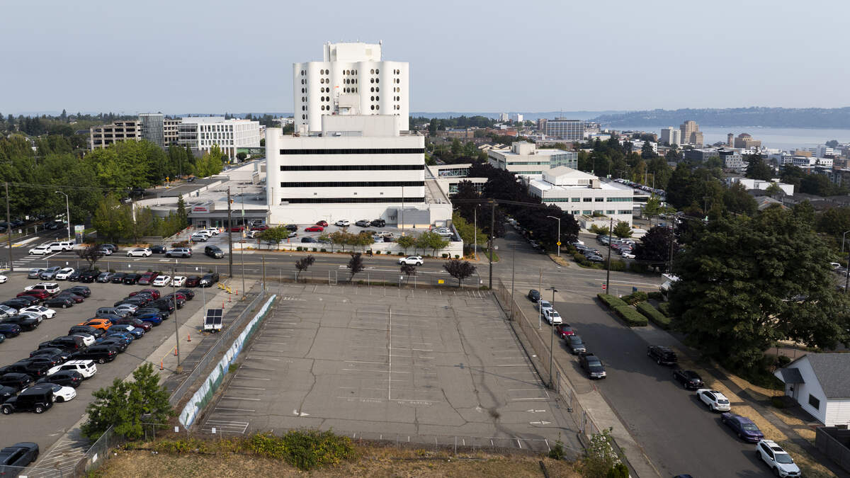Aerial view of a large white multi-story hospital-like building with surrounding parking lots and streets near a waterfront cityscape.