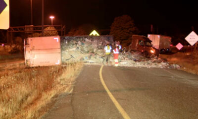Overturned truck blocking a road at night with scattered cargo and two workers in reflective vests inspecting the scene.