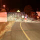 Overturned truck blocking a road at night with scattered cargo and two workers in reflective vests inspecting the scene.
