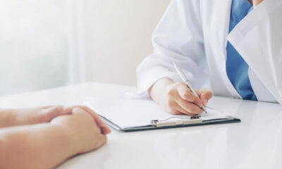Doctor in a white coat writes on a clipboard during a patient consultation, hands resting on the table nearby.