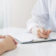 Doctor in a white coat writes on a clipboard during a patient consultation, hands resting on the table nearby.