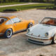 Two vintage Porsche convertibles parked on a gravel driveway with neatly trimmed shrubs in the background.