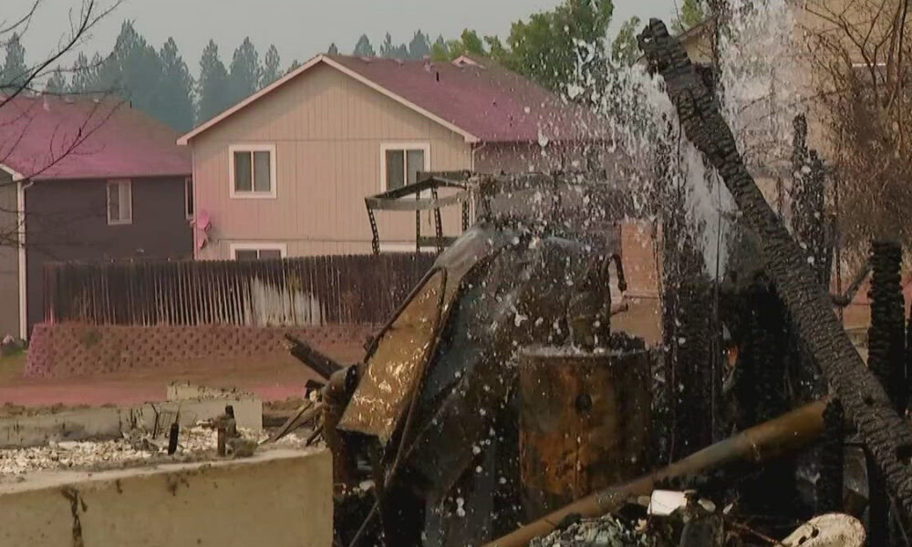 Charred remains of a collapsed structure and debris in a residential yard, with two houses in the background.