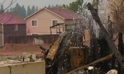 Charred remains of a collapsed structure and debris in a residential yard, with two houses in the background.