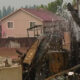 Charred remains of a collapsed structure and debris in a residential yard, with two houses in the background.