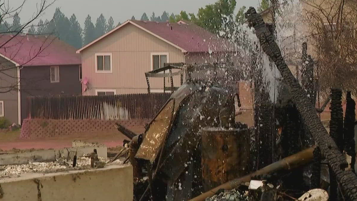 Charred remains of a collapsed structure and debris in a residential yard, with two houses in the background.