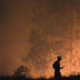 Silhouette of a firefighter wearing a helmet, standing on a small hill and looking toward a raging forest fire with an orange glow behind.