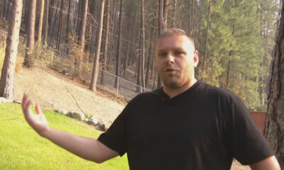 Man in a black T-shirt speaks outdoors with his left arm extended, standing on grass with pine trees and a fence in the background.
